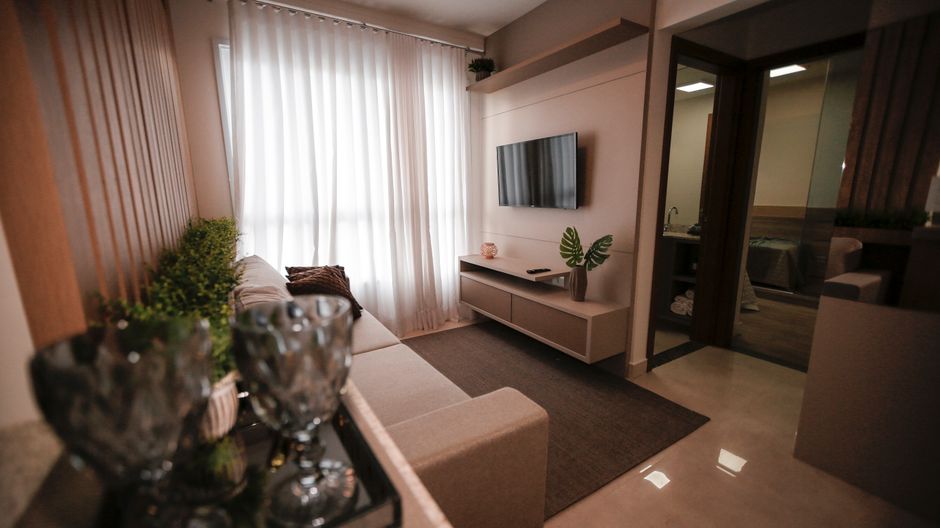 A modern living room with a beige fabric sofa positioned against a wall in front of a large window dressed with white curtains, allowing natural light to illuminate the space. The room features a whit
