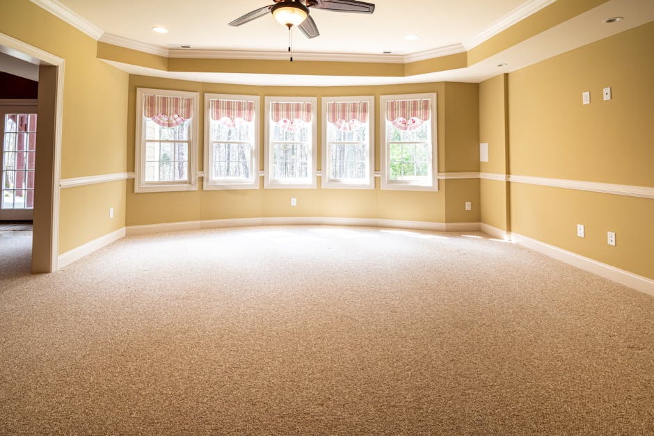 A person operating a yellow commercial carpet cleaning machine on a patterned fabric carpet in a room with warm lighting. The individual holds the device's handle with one hand and presses a control b