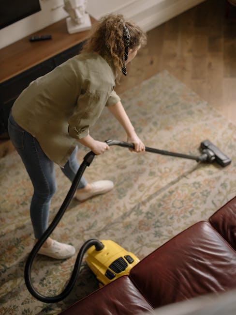 A professional cleaner dressed in a white protective suit, gloves, and face mask using a steam cleaning machine on a beige carpet in a bright living room. The room features light-colored walls, large